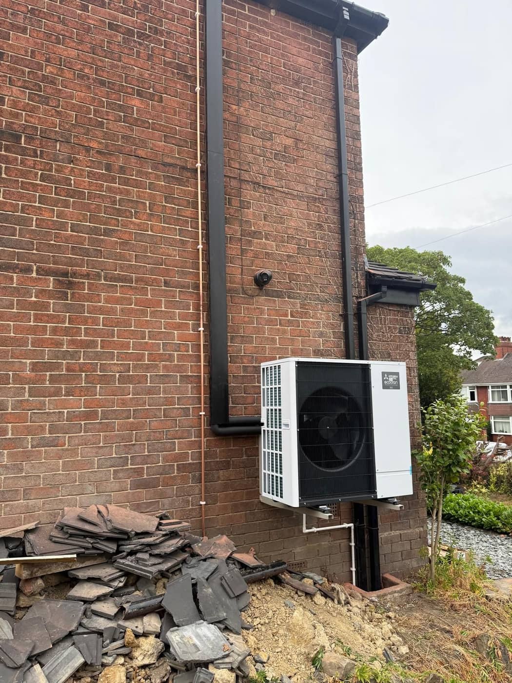 A white air source heat pump on a bracket at the foot of a tall brick gable wall