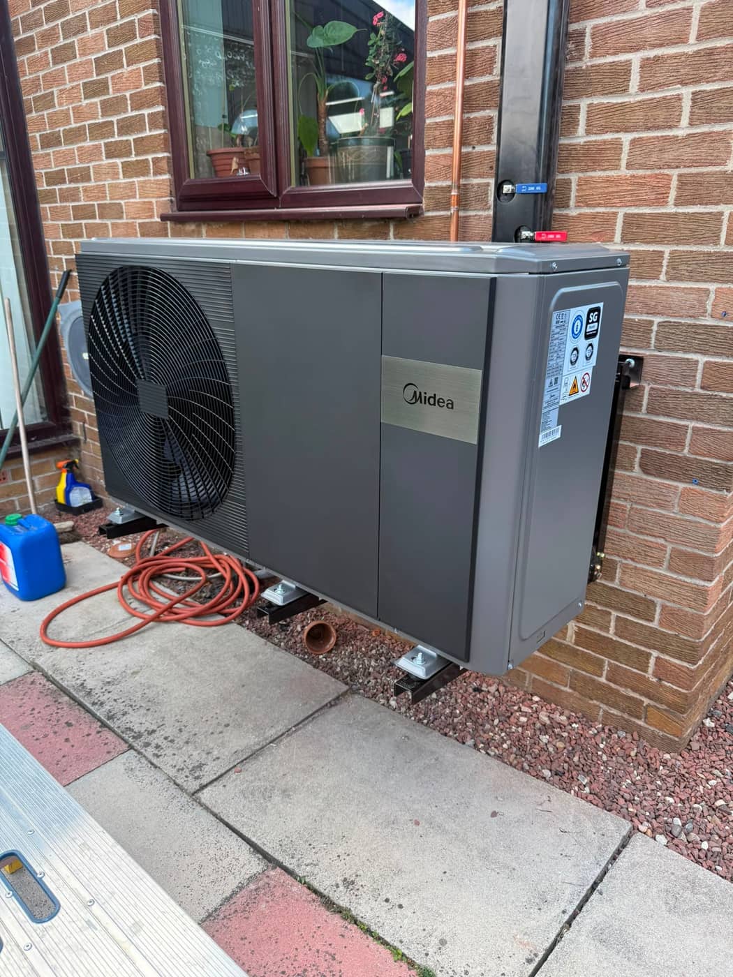 A dark grey air source heat pump on wall brackets against a red brick wall, below a window