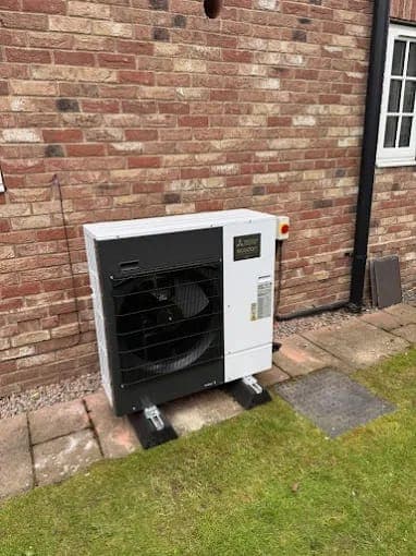 A white heat pump on its feet against a brick wall at the edge of a lawn