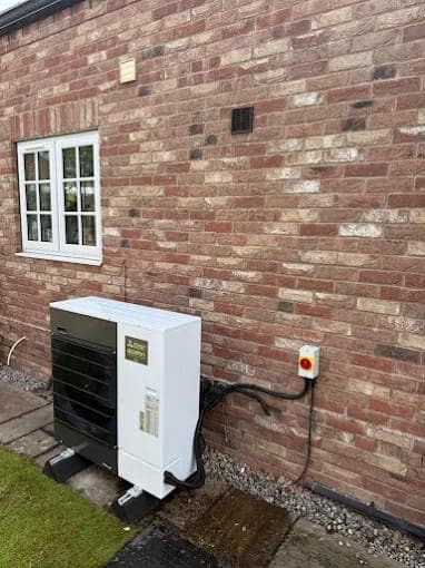 A white heat pump mounted on a brick wall under a cottage window