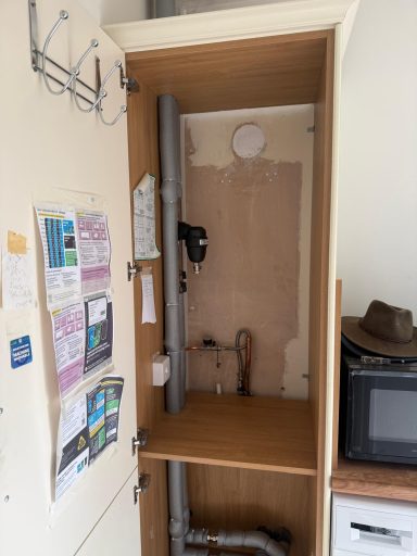 System Boiler Removed Empty cupboard with exposed pipes and bare wall, alongside a small kitchen area.